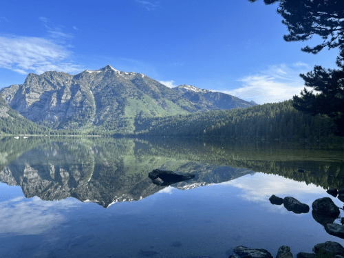 A serene lake reflects majestic mountains and a clear blue sky, surrounded by lush green trees.