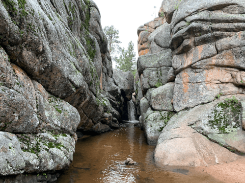 A narrow canyon with towering rock formations and a small waterfall flowing into a shallow pool.