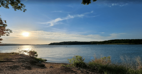 A serene lakeside view at sunset, with trees and gentle waves under a clear blue sky.
