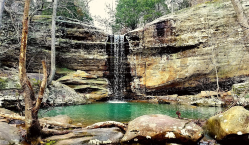 A serene waterfall cascades over rocky cliffs into a tranquil, turquoise pool surrounded by trees and boulders.