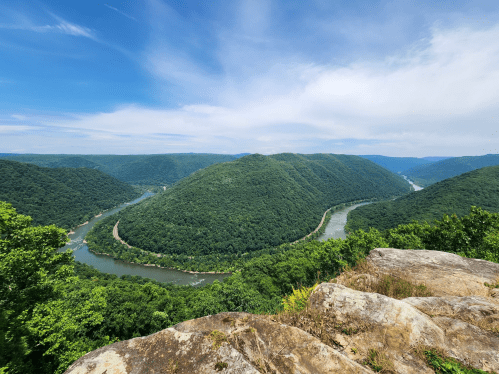 A panoramic view of a winding river surrounded by lush green hills under a blue sky with scattered clouds.