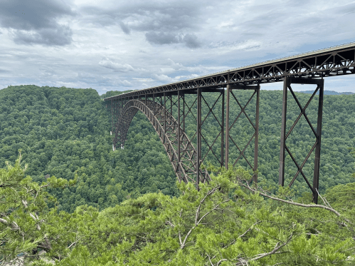 A large metal arch bridge spans a lush green valley under a cloudy sky.