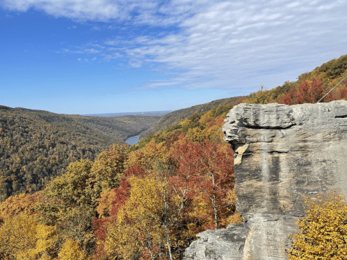A scenic view of a valley with colorful autumn foliage and a river, under a partly cloudy sky.
