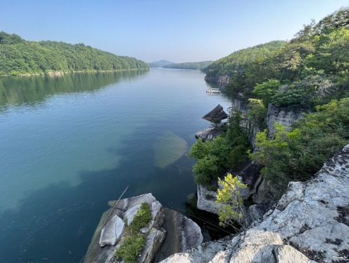A serene river flows between lush green hills, with rocky cliffs and calm waters reflecting the clear blue sky.