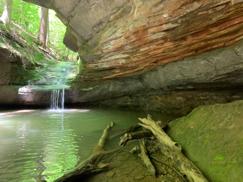 A serene cave with a small waterfall, surrounded by lush greenery and a calm pool of water.