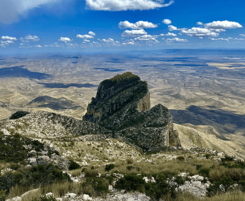 A panoramic view of a rugged mountain peak surrounded by vast, arid landscapes and a blue sky with fluffy clouds.