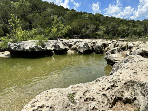 A serene natural scene featuring a clear pool of water surrounded by rocky terrain and lush greenery under a blue sky.