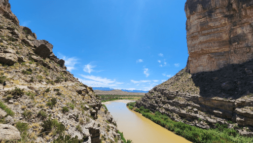 A scenic view of a river winding through rocky cliffs under a clear blue sky. Lush greenery lines the riverbank.
