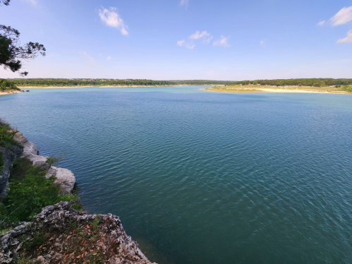 A serene lake surrounded by greenery under a clear blue sky, with gentle waves and rocky shoreline.