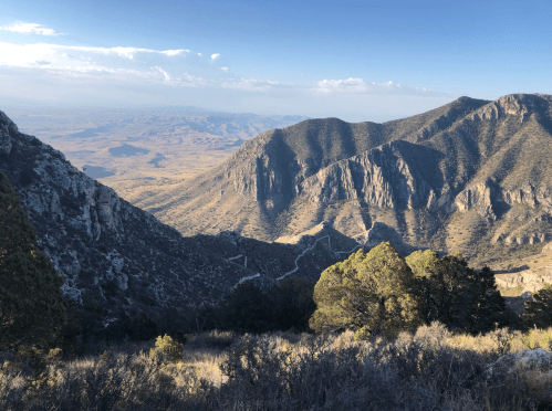 A panoramic view of rugged mountains and valleys under a clear sky, with sparse vegetation in the foreground.