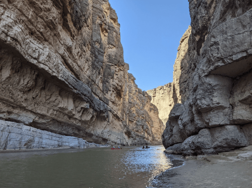 A narrow canyon with steep rock walls and a calm river, with small boats visible in the distance under a clear blue sky.