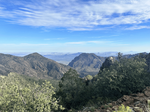 A panoramic view of mountains and valleys under a blue sky with scattered clouds, framed by greenery in the foreground.