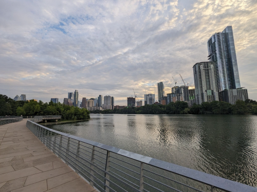 A serene view of a city skyline reflected in a calm river, with clouds overhead and greenery along the shore.