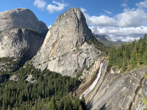 A scenic view of a large granite mountain with a waterfall, surrounded by lush green trees and a blue sky with clouds.