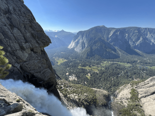 A breathtaking view from a cliff overlooking a waterfall, mountains, and a lush valley under a clear blue sky.