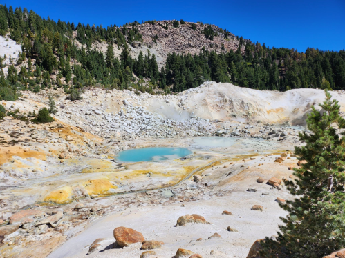 A geothermal area with a turquoise pool surrounded by rocky terrain and pine trees under a clear blue sky.