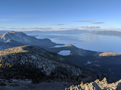 A panoramic view of mountains and a lake under a clear blue sky, with distant hills and water in the background.