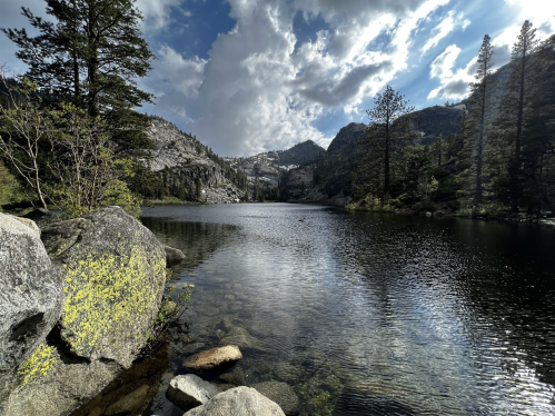 A serene lake surrounded by rocky shores and tall trees, with mountains and clouds in the background.