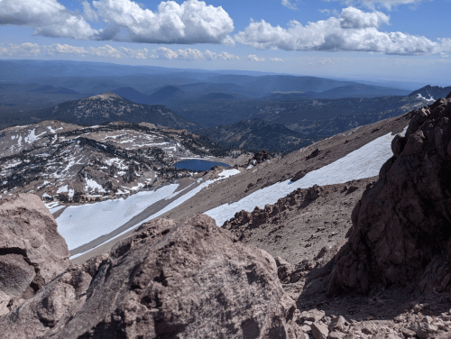 A panoramic view of a mountainous landscape with a lake, rocky terrain, and scattered snow under a cloudy sky.