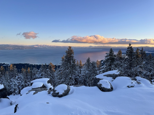 Snow-covered landscape with trees and rocks, overlooking a lake and mountains under a clear blue sky at sunset.