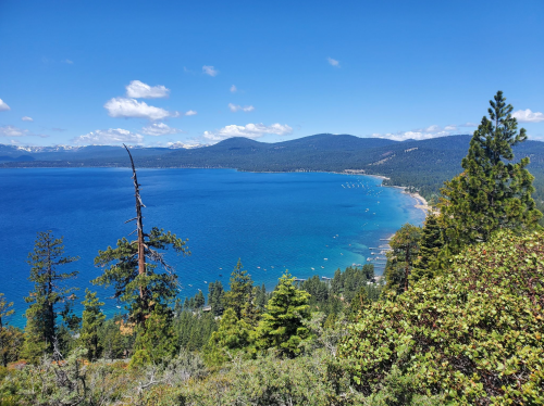 A scenic view of a blue lake surrounded by mountains and trees under a clear sky with a few clouds.