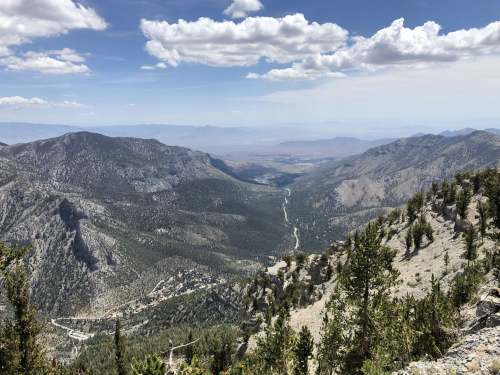 A panoramic view of a mountainous landscape with valleys, trees, and a winding road under a partly cloudy sky.