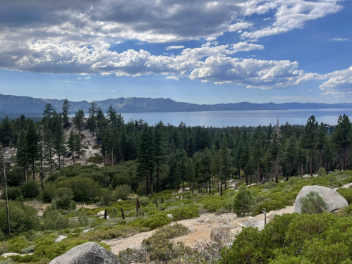 A scenic view of Lake Tahoe surrounded by lush green trees and rocky terrain under a partly cloudy sky.
