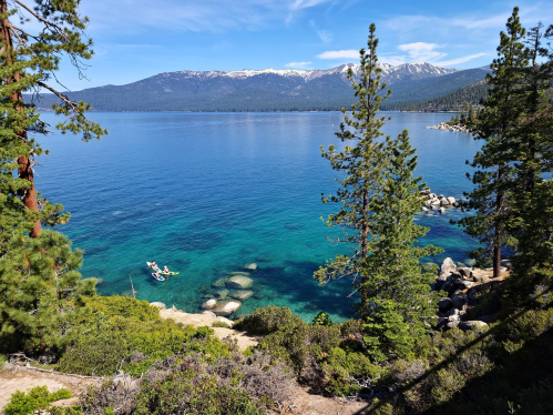 A serene lake scene with clear blue water, rocky shores, and distant mountains, framed by green trees.