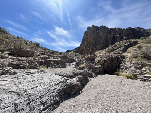 A rocky canyon landscape under a bright blue sky with scattered clouds and sunlight illuminating the terrain.