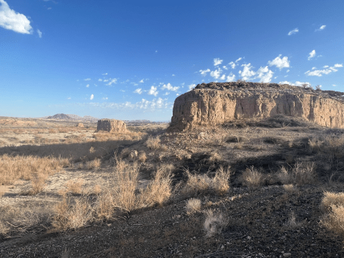 A rocky landscape with dry vegetation under a blue sky, featuring distant hills and scattered clouds.