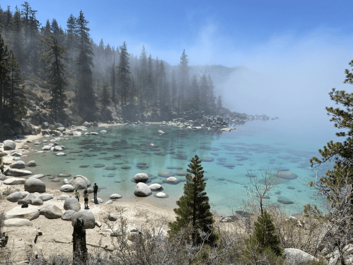 A serene lakeside scene with clear water, smooth rocks, and misty trees in the background. Two people stand on the shore.