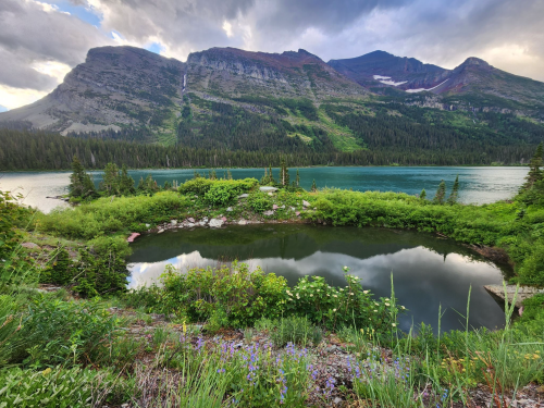A serene landscape featuring a calm lake, lush greenery, and majestic mountains under a cloudy sky.