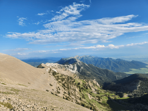 A panoramic view of mountains under a blue sky with scattered clouds, showcasing rugged terrain and lush green valleys.