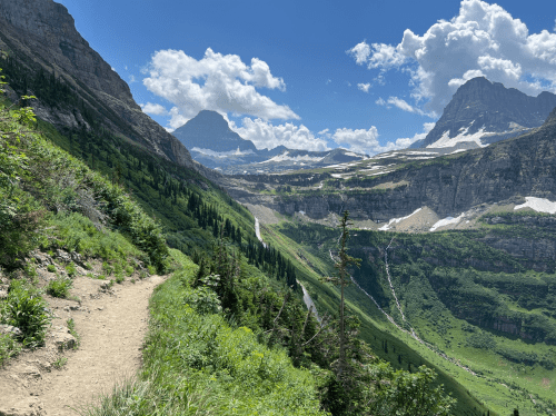 A scenic mountain trail winds through lush greenery, with towering peaks and a blue sky dotted with clouds in the background.