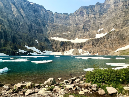 A serene mountain lake surrounded by towering cliffs and scattered icebergs, with greenery in the foreground.