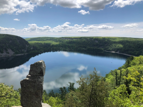 A serene lake surrounded by lush green hills under a partly cloudy sky, with a rock formation in the foreground.