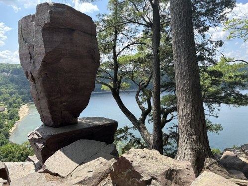 A large rock formation stands on a cliff overlooking a serene lake, surrounded by trees and a clear blue sky.