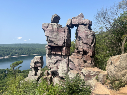 Rock formations stand tall against a clear blue sky, overlooking a serene lake and lush green landscape.