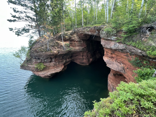 A rocky cliff with lush greenery overhangs a calm, blue-green lake, featuring a cave entrance on the water's edge.