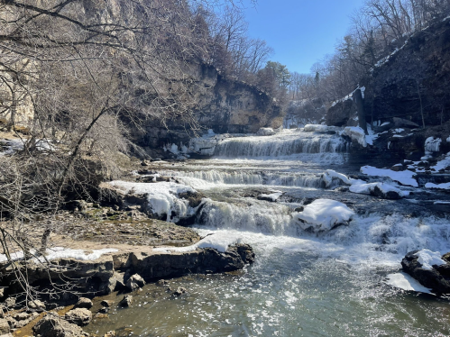 A serene waterfall cascades over rocky terrain, surrounded by bare trees and snow under a clear blue sky.
