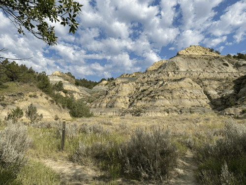 A scenic view of rugged hills under a cloudy sky, with sagebrush and a trail marker in the foreground.