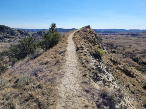 A narrow dirt path winds along a ridge, surrounded by dry, rolling hills under a clear blue sky.