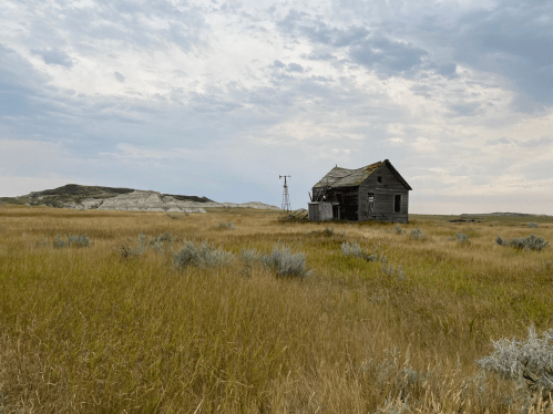 An old, weathered house stands in a grassy field, with a windmill nearby and distant hills under a cloudy sky.