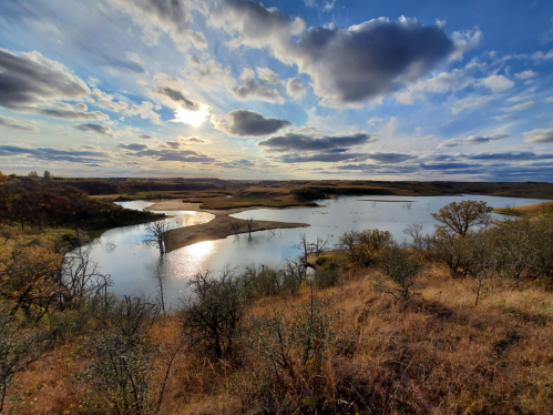 A serene landscape featuring a calm lake, rolling hills, and a dramatic sky with clouds at sunset.