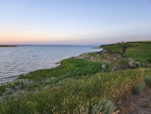 A serene lakeside view at dusk, with lush green grass and gentle waves under a pastel sky.
