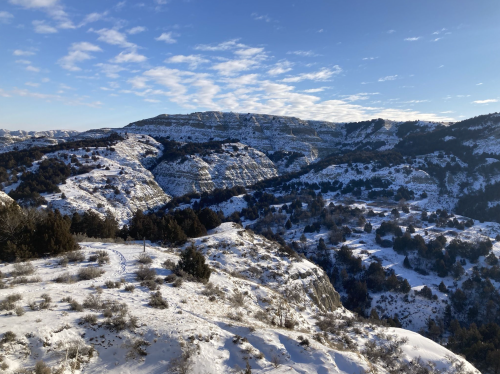 Snow-covered hills and valleys under a blue sky with scattered clouds, showcasing a serene winter landscape.