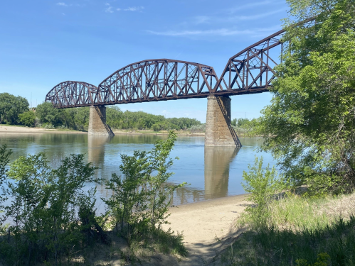 A rusted metal bridge spans a calm river, surrounded by greenery and sandy banks under a clear blue sky.