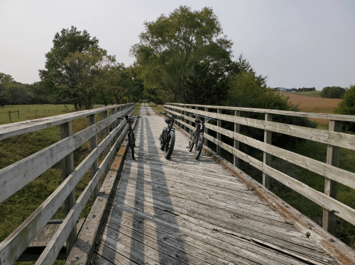 Two bicycles parked on a wooden bridge surrounded by trees and open fields under a clear sky.