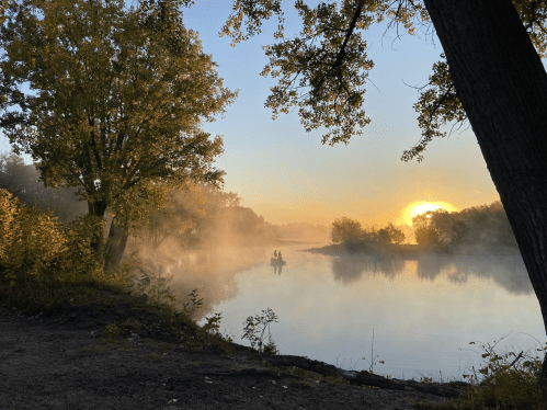 A serene river scene at sunrise, with mist rising and two people in a canoe silhouetted against the golden light.