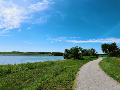 A serene pathway alongside a calm lake, with lush greenery and a clear blue sky above.
